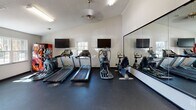 Located at 100 Bermuda Run in Statesboro, Georgia, this single-story community amenity center offers a well-rounded selection of facilities designed to support both active and social lifestyles. The fitness center is a standout feature, thoughtfully divided into distinct workout zones. One area houses a row of cardio equipment — including treadmills, ellipticals, and a stationary bike — positioned in front of floor-to-ceiling mirrors and wall-mounted televisions, with natural light streaming in through exterior glass doors. A separate strength training zone features a comprehensive dumbbell rack, multi-use cable machine, and additional resistance equipment, all set on durable rubber flooring beneath vaulted ceilings with ceiling fans. The business center provides a quiet, functional workspace with a long desk running along a deep teal-painted wall, three computer workstations, brass wall sconces, and framed abstract artwork overhead. The social lounge is an inviting open-plan space finished with wood-look flooring, bold dark trim, and a distinctive tiled bar-height island at its center. Hanging bubble chairs and colorful drapery add a playful, modern character to the room. A pool table and open seating areas complete this communal gathering space, making it ideal for relaxation and recreation.