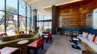 This expansive lobby is a masterfully designed space that blends contemporary architecture with bold, eclectic interiors. Upon entry, visitors are greeted by a striking dark marble reception desk positioned beneath towering vertical wood slat screens that serve as dramatic architectural dividers throughout the space. The double-height ceilings amplify the sense of openness, while floor-to-ceiling glass walls along the perimeter flood the interior with natural light and frame views of the surrounding urban landscape.

The main lounge area is anchored by a sculptural chandelier of cascading metallic branches, suspended above a curated arrangement of curved sage-green seating, vibrant red accent chairs, and dark wood tables arranged atop a graphic navy patterned rug. Teal banquette seating with dark marble tables offers an intimate setting framed by warm wood slat partitions.

A glazed café area features geometric-patterned tile flooring in slate blue and white, complemented by warm wood furnishings and panoramic city views. Secondary corridors showcase textured stone wall cladding and large-format tile floors, leading to glass-enclosed meeting or lounge spaces. A utility alcove features richly tiled amber walls and bold geometric floor patterns. Throughout, the design balances warmth, texture, and contemporary elegance.
