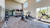 Located at 510 Morning Oaks Dr in Westfield, Indiana, this fitness center is a thoughtfully designed, single-floor facility offering two dedicated exercise rooms and an adjoining patio. The main exercise area impresses with soaring double-height ceilings and an abundance of natural light streaming through large windows and glass doors, creating an open and energizing atmosphere. The space is outfitted with a full suite of cardio equipment, including treadmills, ellipticals, and stationary bikes, all arranged across durable, patterned carpet tile flooring. A dedicated strength and functional training zone features a multi-use cable machine, weight benches, and resistance equipment. Floor-to-ceiling mirrors along one wall visually expand the space and provide practical feedback during workouts, while wall-mounted flat-screen televisions add entertainment options. A glass-partitioned studio room offers a versatile space for group fitness or flexibility training, complete with stability balls and additional equipment. A built-in water fountain station with tile surround provides convenient hydration access. Accent walls in deep teal and dark charcoal add contemporary visual interest throughout. The connected patio provides an outdoor extension of the facility, with views of the surrounding greenery, enhancing the overall wellness environment.