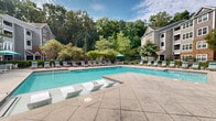Located at Avonlea Place in Woodstock, Georgia, this outdoor amenity area offers a well-appointed resort-style retreat designed for relaxation and recreation. At the heart of the space is a generously sized swimming pool with crystal-clear water, featuring in-pool lounge chairs and a gradual entry that invites leisurely enjoyment. The pool deck is finished with textured concrete and natural flagstone pavers, providing ample space for sunbathing on the numerous cushioned chaise lounges arranged throughout. Colorful, manicured landscaping with flowering plants, sculpted shrubs, and mature trees frames the entire area, creating a lush, private atmosphere. A fenced perimeter ensures a secure environment while preserving the open, airy feel. Adjacent to the pool is a craftsman-style clubhouse and covered pavilion featuring an outdoor seating area with dining furniture and a wall-mounted television, making it ideal for gatherings. The structure incorporates brick and board-and-batten siding with a pitched roof, adding architectural character. Teal umbrellas throughout the deck provide shade and a cohesive aesthetic. The backdrop of towering trees and multi-story residential buildings surrounding the space enhances the sense of a secluded, community-centered oasis in the heart of Woodstock, GA.