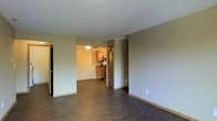 This single-floor space offers a straightforward, functional layout centered around an open living area that flows directly into the kitchen. The living space features dark hardwood-style flooring that contrasts with the neutral beige walls, creating a clean and understated aesthetic. A textured popcorn ceiling runs throughout, adding a classic touch to the interior.

The kitchen is visible from the main living area and is equipped with warm medium-toned wood cabinetry, stainless steel appliances including a range and microwave, and a dark countertop. A ceiling fan with a light fixture is installed in the kitchen area, providing both ventilation and overhead lighting.

A louvered bi-fold door adjacent to the kitchen likely conceals a closet or utility area, offering practical storage within the unit. On the opposite side of the living space, a door with a mirrored surface—likely a closet—adds a reflective element that helps enhance the sense of openness in the room.

The overall ambiance is modest and neutral, designed for practical everyday living with a simple, no-frills interior that provides a versatile blank canvas for personalization.