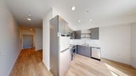 Located at 3725 N 27th St in Tacoma, Washington, this single-floor unit offers a clean, modern aesthetic throughout. The open-concept layout is anchored by a well-appointed kitchen featuring sleek gray cabinetry with bar-pull hardware, white quartz countertops, and a full suite of stainless steel appliances including a refrigerator, electric range, over-the-range microwave, and dishwasher. Light wood-tone flooring runs continuously throughout the space, enhancing the bright and airy feel. Large sliding glass doors in the main living area flood the space with natural light and provide direct access to a private balcony, where scenic outdoor views can be enjoyed. The bathroom is thoughtfully designed with dark wood cabinetry, a white countertop vanity, a wall-mounted storage cabinet, and a walk-in shower with grab bars. A stacked washer and dryer unit is conveniently integrated within the bathroom area, offering in-unit laundry. The overall design is contemporary and functional, with crisp white walls, recessed lighting, and a cohesive neutral palette that lends the space a clean, move-in-ready appeal.