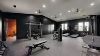 Located at 100 Foxborough Way in Richmond, Kentucky, this fitness center offers a comprehensive exercise facility divided into two distinct exercise rooms, all on a single level. The main exercise room is finished with bold matte-black walls and vaulted ceilings accented by recessed lighting and ceiling fans, creating a focused, high-energy atmosphere. Durable speckled rubber flooring spans the space, providing both safety and durability underfoot. This room is outfitted with professional-grade strength equipment including a power rack with bench press, cable machines, a functional trainer, and a variety of selectorized machines. A row of modern treadmills and an elliptical trainer with built-in screens lines the window wall, allowing natural light and outdoor views during cardio sessions. The second exercise room features a warm, earthy orange accent wall that contrasts with the dark trim and large multi-pane windows, which flood the space with natural light and offer views of the surrounding neighborhood. This room is dedicated to functional and group fitness, housing an Echelon spin bike, medicine ball and resistance band storage, stability balls, and an interactive fitness screen. Together, the two rooms provide a well-rounded fitness experience suited for a wide range of training styles.