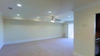 This single-floor space presents a generously sized main living area designed with a clean, neutral aesthetic. The room is finished with light beige wall-to-wall carpeting that extends throughout, contributing to a warm and understated atmosphere. Crisp white walls are complemented by white crown molding and baseboards, lending the interior a polished, traditional character.

The ceiling features elegant tray detailing, adding architectural interest and a sense of depth overhead. Recessed lighting is distributed evenly across the ceiling, providing ample ambient illumination throughout the space. A centrally positioned ceiling fan with a built-in light fixture offers both practical airflow and supplementary lighting.

Natural light enters the room through a large window dressed with vertical blinds, which can be adjusted to control light levels and privacy. A floor-mounted vent is visible along the exterior wall, indicating a central heating and cooling system. The overall layout of the room is open and unobstructed, offering flexibility for a variety of furniture arrangements. The space conveys a well-maintained, move-in-ready condition with a neutral palette suitable for a range of interior design styles.