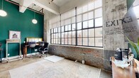 This distinctive industrial loft blends raw architectural character with thoughtfully curated modern design. The main living and workspace area is the centerpiece of the floor plan, boasting soaring ceilings that amplify the sense of openness and volume. Expansive, multi-pane factory-style windows with black steel frames stretch nearly the full height of the exterior wall, flooding the interior with abundant natural light and offering views of the surrounding urban streetscape. Exposed brick walls — some in their natural reddish tone, others painted a soft whitewash — run alongside concrete columns and weathered plaster surfaces, lending authentic industrial character throughout. Bold teal-painted accent walls provide a vibrant contrast to the raw textures, and polished concrete floors tie the aesthetic together seamlessly. The living and office area comfortably accommodates both a lounge setting and a dedicated workspace, with warm pendant globe lighting adding a soft glow overhead. The space also includes a full bathroom finished in a clean, minimalist style, featuring crisp white walls and cabinetry, a glass-enclosed walk-in shower, and polished concrete flooring that echoes the industrial palette found throughout. Overall, the space delivers a compelling fusion of urban grit and contemporary refinement.