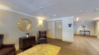 10 Landing Lane presents a thoughtfully designed lobby level that combines classical elegance with practical functionality. The main entry corridor leads to a generously proportioned mail room lined on both sides with gold-toned mailboxes, illuminated by recessed linear lighting in the ceiling and finished with wood-look tile flooring. Glass doors at the far end of the corridor invite natural light deep into the space, creating a bright and open feel.

The adjacent lounge area exudes a refined, residential warmth. Rich hardwood flooring, crisp white crown molding, and subtly textured wallcoverings set a sophisticated backdrop. Tasteful furnishings, including tufted leather wingback chairs and dark wood console cabinets, are arranged throughout, while a gold sunburst mirror serves as a striking decorative focal point. Warm wall sconces cast a soft, inviting glow, complementing the recessed ceiling lights overhead. Framed artwork adds a curated, gallery-like quality to the space.

Together, these areas establish a polished and welcoming first impression, balancing the practical needs of a multi-residence building with the comfort and aesthetic sensibility of a well-appointed interior. The consistent use of warm tones, quality materials, and classical architectural details throughout the lobby creates a cohesive and distinguished environment at 10 Landing Lane.