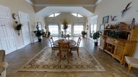 This inviting single-floor space makes an immediate impression with its dramatic vaulted ceilings that rise to a striking peak, creating a grand sense of openness and volume. Large clerestory windows positioned high along the roofline flood the interior with natural light, complementing the warm, neutral palette of the walls and tile flooring. The main gathering area centers around a classic round wooden dining table surrounded by cross-back chairs, all set atop an ornate traditional area rug with intricate floral detailing that anchors the space with warmth and elegance. Recessed lighting supplements the abundant daylight, ensuring the room remains bright and welcoming at all hours. A row of generously sized windows along the far wall, dressed with tailored roller shades, frames views to the outdoors and enhances the airy atmosphere. Lush potted greenery is thoughtfully placed throughout, adding life and color to the neutral tones. A handsome wooden hutch or display cabinet along one wall provides both storage and a decorative focal point. The open floor plan allows for easy movement throughout, with a seamless blend of functional and decorative elements that convey a comfortable, well-appointed, and timeless aesthetic.
