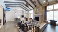Located at 1680 Bluewater Way in Charleston, South Carolina, this single-story space makes an immediate impression with its striking architectural design and refined interior aesthetic. The main living area commands attention with soaring vaulted ceilings adorned with bold exposed dark beams, creating a dramatic sense of height and openness. A series of geometric pendant light fixtures and delicate glass globe pendants hang gracefully from the ceiling, casting warm, layered lighting throughout the space. A sleek, floor-to-ceiling fireplace serves as a natural focal point, framed by smooth stone surround and complemented by curated wall art above. The flooring features wide-plank hardwood in a warm neutral tone that flows seamlessly throughout, anchoring the space with a sense of continuity. The open layout connects the living area to what appears to be a bar or refreshment station, complete with pendant lighting and counter seating, making it well-suited for entertaining. Large windows invite generous natural light, enhancing the airy, welcoming ambiance. The overall design blends contemporary and transitional styles, with neutral tones, layered textures, and carefully chosen furnishings that reflect a sophisticated and cohesive interior vision.