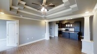This single-floor home showcases a thoughtfully designed open-concept layout that combines comfort with refined architectural character. The living area immediately draws attention with its multi-tiered tray ceiling, accented by crisp crown molding and a centrally mounted ceiling fan that adds both function and visual appeal. Neutral gray walls complement the warm, light-toned hardwood flooring that flows throughout the space, creating a cohesive and inviting atmosphere.

A decorative white column serves as an elegant architectural divider between the living and kitchen areas, adding a classic touch without interrupting the open flow of the floor plan. The kitchen is a standout feature, appointed with rich dark cabinetry, stone countertops, a tasteful tile backsplash, and pendant lighting that hangs above the counter space. Stainless steel appliances and ample cabinetry provide both style and practicality.

The overall design blends traditional and contemporary elements, with careful attention to detail evident in the millwork, column accents, and layered ceiling design. The generous proportions of the main living space and the well-appointed kitchen make this home ideal for both everyday living and entertaining. The neutral color palette throughout ensures versatility and a timeless aesthetic appeal.