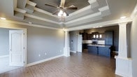 This single-floor home showcases a thoughtfully designed open-concept layout that combines comfort with refined architectural character. The living area immediately draws attention with its multi-tiered tray ceiling, accented by crisp crown molding and a centrally mounted ceiling fan that adds both function and visual appeal. Neutral gray walls complement the warm, light-toned hardwood flooring that flows throughout the space, creating a cohesive and inviting atmosphere.

A decorative white column serves as an elegant architectural divider between the living and kitchen areas, adding a classic touch without interrupting the open flow of the floor plan. The kitchen is a standout feature, appointed with rich dark cabinetry, stone countertops, a tasteful tile backsplash, and pendant lighting that hangs above the counter space. Stainless steel appliances and ample cabinetry provide both style and practicality.

The overall design blends traditional and contemporary elements, with careful attention to detail evident in the millwork, column accents, and layered ceiling design. The generous proportions of the main living space and the well-appointed kitchen make this home ideal for both everyday living and entertaining. The neutral color palette throughout ensures versatility and a timeless aesthetic appeal.