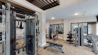 This fitness center offers a comprehensive and thoughtfully designed workout environment within a single open floor. The space is defined by its striking architectural ceiling treatment, featuring dark wood slat panels and exposed beam-style elements that add visual depth and a contemporary industrial character against the crisp white walls and columns. Patterned, neutral-toned carpet flooring runs throughout, providing both comfort and durability underfoot.

The cardio zone is well-appointed with multiple treadmills, elliptical trainers, upright bikes, and a stair climber, all positioned to face large floor-to-ceiling windows that frame views of the outdoor pool area and surrounding grounds, allowing ample natural light to fill the space. Wall-mounted flat-screen televisions are strategically placed throughout to enhance the workout experience.

The strength training area features a multi-functional cable machine, a weight-assisted resistance system, adjustable benches, and a dedicated free weight section with a fully stocked dumbbell rack. Large mirrors line portions of the walls, visually expanding the space and allowing members to monitor their form. The overall ambiance is modern, clean, and energizing, creating a functional yet aesthetically refined environment suited for a wide range of fitness routines.