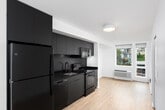 This single-floor unit showcases a clean, contemporary design defined by crisp white walls, light hardwood-style flooring, and a cohesive modern aesthetic throughout. The kitchen is a standout feature, outfitted with floor-to-ceiling matte dark grey cabinetry, dark stone countertops, under-cabinet lighting, and a full suite of black and stainless steel appliances including an electric range, built-in microwave, dishwasher, and refrigerator. A textured grey backsplash and matte black faucet complete the polished look, while a window provides natural light to the cooking area.

The living area is bright and open, with a large floor-to-ceiling window offering street-level views, allowing natural light to fill the space. A wall-mounted thermostat and recessed lighting add functional detail throughout.

The bathroom maintains the unit's modern character with a white tub-shower combination, matte black fixtures and grab bar, tile flooring in a warm grey tone, and a wall-mounted vanity. Adjacent to the main living area, a dedicated walk-in closet features a clothing rod and ample wall space for storage. The unit's efficient layout, visible in the floorplan scan, ensures a logical and comfortable flow between all areas.