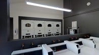 This single-floor laundry facility is designed with functionality and efficiency in mind, featuring a clean, utilitarian layout suited for high-volume use. The space is finished with a two-tone color scheme, combining crisp white equipment against dark gray walls, giving the room a modern and organized appearance.

Along the back wall, a row of commercial-grade front-loading dryers is mounted on raised pedestals, providing convenient access and maximizing the use of vertical space. Each dryer unit features a large porthole-style door and a control panel at the base, indicating coin or card-operated functionality. The units appear to be well-maintained and uniform in design.

In the foreground, a lineup of top-loading commercial washers is positioned in a parallel row, each equipped with digital control panels and visible water supply connections, reflecting a professional-grade setup. Instructional signage is posted on the wall, guiding users through proper machine operation.

Overhead, bright fluorescent tube lighting runs the length of the ceiling, ensuring the entire space is well-lit and easy to navigate. The overall ambiance is practical and no-frills, prioritizing operational efficiency and cleanliness.