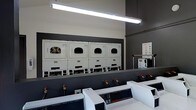 This single-floor laundry facility is designed with functionality and efficiency in mind, featuring a clean, utilitarian layout suited for high-volume use. The space is finished with a two-tone color scheme, combining crisp white equipment against dark gray walls, giving the room a modern and organized appearance.

Along the back wall, a row of commercial-grade front-loading dryers is mounted on raised pedestals, providing convenient access and maximizing the use of vertical space. Each dryer unit features a large porthole-style door and a control panel at the base, indicating coin or card-operated functionality. The units appear to be well-maintained and uniform in design.

In the foreground, a lineup of top-loading commercial washers is positioned in a parallel row, each equipped with digital control panels and visible water supply connections, reflecting a professional-grade setup. Instructional signage is posted on the wall, guiding users through proper machine operation.

Overhead, bright fluorescent tube lighting runs the length of the ceiling, ensuring the entire space is well-lit and easy to navigate. The overall ambiance is practical and no-frills, prioritizing operational efficiency and cleanliness.