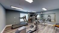 This single-level fitness room offers a practical and organized space designed to support a range of cardiovascular and light strength-training activities. The room is finished with durable wood-look flooring throughout, providing a clean and easy-to-maintain surface that complements the cool blue-gray painted walls. Recessed fluorescent panel lighting illuminates the space evenly, ensuring visibility across all areas of the room.

The cardio equipment selection includes a treadmill, elliptical trainer, stationary recumbent bike, and an additional stepping machine, all positioned to allow comfortable use without overcrowding. A dumbbell rack with a wall-mounted television is situated near a window fitted with horizontal blinds, allowing natural light to filter in while maintaining privacy. A large wall mirror spans one side of the room, visually expanding the space and allowing users to monitor their form during exercise. A wall clock is mounted nearby for easy time-tracking.

Seating is available via a chair positioned near the equipment, and a hand sanitizer dispenser is mounted on the wall for convenience. An interior window and doorway connect the fitness room to an adjacent meeting or activity room, suggesting this space is part of a larger communal facility.