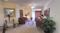 This single-floor residence centers around a welcoming living room designed for both comfort and functionality. The space is finished with plush wall-to-wall carpeting in a neutral tone, providing a soft foundation that complements the warm, earthy color palette throughout. Rich burgundy upholstered seating—including a sofa and coordinating armchairs—anchors the room, arranged thoughtfully around a glass-top coffee table with a dark metal frame. A ceiling fan with an integrated light fixture is centrally positioned overhead, offering both ambient illumination and air circulation.

Natural light enters through a sliding glass door dressed with deep red curtains and a transom window above, hinting at outdoor access. The walls are painted a soft, warm yellow-white, adorned with framed artwork and decorative wall accents that add visual interest without overwhelming the space. Table lamps with dark shades provide additional warm lighting throughout. Lush indoor plants placed near the entryway and seating area introduce a refreshing natural element. A small bistro-style table and chairs are tucked to one side, adding a versatile spot for casual use. Overall, the space exudes a cozy, well-appointed atmosphere.