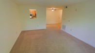 This single-story space features an open-concept layout that connects the living area to an adjacent entry zone, creating a sense of continuity throughout the main living space. The living area is finished with wall-to-wall neutral beige carpeting, contributing to a warm and understated aesthetic. A ceiling fan with a built-in light fixture is centrally mounted, providing both air circulation and ambient lighting.

A notable architectural detail is the pass-through window that connects the kitchen to the living area, allowing for easy communication and light flow between the two spaces. The kitchen, visible through the pass-through, features warm wood-toned cabinetry and includes appliances such as a microwave, offering a functional and efficient layout.

The entry area is defined by a white paneled door and is illuminated by the ceiling fan fixture, creating a welcoming transition point. Floor vents are positioned throughout for climate control, and ample wall outlets are distributed conveniently. The overall color palette—soft greens, whites, and warm neutrals—lends the space a clean, move-in-ready feel suitable for a variety of furnishing styles.