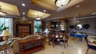 This sophisticated single-floor common space is designed with a classic, upscale aesthetic that blends comfort and refined elegance throughout. The room's most striking architectural feature is its layered coffered ceiling, finished with ornate gold-toned detailing and crown molding, creating a sense of grandeur. Recessed lighting and pendant fixtures illuminate the space warmly, complementing the rich earth-tone walls.

The main gathering area features multiple distinct seating zones, including a circular dining table surrounded by dark wood chairs with navy upholstered seats, a pair of ornate high-back accent chairs positioned near large windows, and a cushioned settee flanked by traditional wooden furniture including a substantial bookcase-style cabinet. Travertine tile flooring and a decorative area rug add texture and warmth underfoot.

A wet bar area, accessible from the main room, includes a curved granite countertop with bar seating, dark wood cabinetry with glass-front uppers, and pendant lighting overhead. Large windows and glass-paned doors along one wall flood the space with natural light and provide direct access to an outdoor patio area, where views of tropical greenery and a pool are visible.