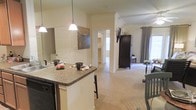 This single-floor residence showcases an open-concept design that connects the kitchen, dining, and living areas into one cohesive, flowing space. The kitchen features a peninsula with granite countertops and bar seating, ideal for casual dining or entertaining. Pendant lighting hangs above the counter, adding a decorative touch while providing functional task lighting. Crown molding along the ceilings adds a refined architectural detail throughout the space. The flooring transitions from hard surface near the kitchen to plush carpeting in the living and adjacent areas, contributing to a warm and comfortable ambiance. Neutral, cream-toned walls allow natural light to brighten the interior, complemented by a window dressed with curtains in the living area. A dark wood armoire serves as a notable furniture piece, adding contrast against the light palette. A hallway visible from the kitchen leads to additional rooms, hinting at a well-organized layout. Decorative elements, including wall art and floral accents, enhance the inviting atmosphere of this thoughtfully appointed space.