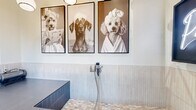 This 70-square-foot pet grooming room is a thoughtfully designed, single-floor amenity space dedicated entirely to pet care. The centerpiece of the room is a raised, built-in dog wash station clad in slate-blue rectangular tiles, complete with an integrated handheld spray attachment and a mosaic-tiled surface for pets to stand on. A tiled step on one side provides easy access for pets of all sizes. A professional-grade pet dryer is stationed at the edge of the wash area, and a rolling utility cart nearby offers convenient storage for grooming supplies and accessories. The walls feature vertical shiplap-style wainscoting in a light gray tone, creating a clean and modern aesthetic. Overhead, globe pendant lights provide warm, even illumination throughout the space. The walls are adorned with large-format black-and-white photographic prints of dogs, adding a charming and lighthearted character to the room. A standout decorative element is a glowing neon "Paw Day" sign mounted in a black frame, serving as a fun focal point. Decorative chrome sphere wall accents add a polished, contemporary touch. The concrete floor completes the practical yet stylish design of this well-equipped pet grooming facility.