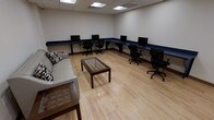 This single-floor business center offers a functional and organized workspace designed to meet a variety of professional needs. The room features a wraparound navy blue desk along two walls, lined with multiple computer workstations, each paired with an ergonomic black office chair, providing ample space for focused work or research. The light wood laminate flooring adds a clean, modern aesthetic that brightens the overall atmosphere. Overhead, recessed fluorescent ceiling tiles provide consistent, even lighting throughout the space. A comfortable light-colored sofa with decorative pillows is positioned along one wall, accompanied by a wooden coffee table at the center of the room, creating a casual seating area for informal meetings or relaxation between work sessions. The neutral white walls keep the environment feeling open and distraction-free. The layout balances dedicated workstation areas with a lounge-style seating zone, making the space versatile for both individual productivity and collaborative use. Overall, the business center presents a tidy, well-maintained environment suited for guests or professionals requiring reliable access to computer resources and a comfortable place to conduct business.