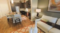 This single-story space showcases an open-concept layout that seamlessly connects the kitchen and living area, creating a cohesive and functional environment. The kitchen is outfitted with rich, dark espresso cabinetry that extends to the upper walls, providing ample storage. Stainless steel appliances, including a refrigerator and built-in microwave, complement the modern aesthetic. A double sink with a sleek faucet sits beneath the upper cabinets, and a breakfast bar with tall seating extends from the kitchen counter, offering a casual dining option.

Warm hardwood flooring flows throughout the space, unifying the kitchen and living areas. The living area is anchored by a plush white armchair and accented with a decorative floor lamp and side table. Bold orange-and-white patterned area rugs add visual interest and warmth to both zones. Neutral, earthy wall tones tie the space together, creating a welcoming ambiance. The open layout maximizes the sense of space, making the interior feel airy and well-organized, ideal for both everyday living and casual entertaining.