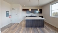 Located at 1185 Shelburne Road in Burlington, Vermont, this thoughtfully designed single-floor residence offers a clean, modern aesthetic throughout. The open-concept living and kitchen area is anchored by light-toned wood-style flooring that flows seamlessly throughout the main space. The kitchen is a standout feature, boasting rich dark cabinetry, white quartz countertops, a large center island with pendant lighting, stainless steel appliances including a dishwasher and microwave, and a double-basin sink. A wall-mounted mini-split unit provides efficient climate control in the living area. The living room is bright and airy, with a sliding glass door leading to a private balcony that offers expansive outdoor views of the surrounding Burlington landscape. Two carpeted bedrooms provide comfortable retreats, each benefiting from natural light through well-placed windows. The primary bedroom includes a dedicated closet and direct access to a full bathroom. Both bathrooms are finished with dark wood vanities, white countertops, large mirrors, and tile flooring, with accessibility grab bars thoughtfully incorporated. The overall ambiance is contemporary, clean, and functional, making this a well-appointed residence in a convenient Burlington location.