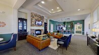 This well-appointed single-floor community clubhouse offers a thoughtfully designed gathering space that balances elegance with functionality. The centerpiece of the main lounge is a dramatic floor-to-ceiling stacked-stone fireplace, topped with a wall-mounted television and flanked by dark wood cabinetry. Comfortable cognac leather sofas and deep navy accent chairs are arranged around a glass coffee table, creating an inviting seating area suited for relaxed socializing. Tray ceilings with recessed lighting add architectural depth and a warm ambiance throughout the lounge. Adjacent to the main seating area, a sunlit dining nook features large windows dressed with patterned drapery, a round table, and navy upholstered chairs beneath a classic chandelier. The space transitions seamlessly into a kitchen and bar area finished in rich teal tones, where dark espresso cabinetry, mosaic tile backsplash, granite countertops, and stainless steel appliances are complemented by pendant lighting and leather-upholstered bar stools. A games room rounds out the amenities, offering a billiards table beneath a statement pendant light, with ample natural light filtering through large windows. Crown molding, coordinated green wainscoting, and tile flooring throughout tie the entire space together cohesively.