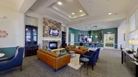 This well-appointed single-floor community clubhouse offers a thoughtfully designed gathering space that balances elegance with functionality. The centerpiece of the main lounge is a dramatic floor-to-ceiling stacked-stone fireplace, topped with a wall-mounted television and flanked by dark wood cabinetry. Comfortable cognac leather sofas and deep navy accent chairs are arranged around a glass coffee table, creating an inviting seating area suited for relaxed socializing. Tray ceilings with recessed lighting add architectural depth and a warm ambiance throughout the lounge. Adjacent to the main seating area, a sunlit dining nook features large windows dressed with patterned drapery, a round table, and navy upholstered chairs beneath a classic chandelier. The space transitions seamlessly into a kitchen and bar area finished in rich teal tones, where dark espresso cabinetry, mosaic tile backsplash, granite countertops, and stainless steel appliances are complemented by pendant lighting and leather-upholstered bar stools. A games room rounds out the amenities, offering a billiards table beneath a statement pendant light, with ample natural light filtering through large windows. Crown molding, coordinated green wainscoting, and tile flooring throughout tie the entire space together cohesively.