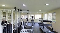 This fitness center offers a thoughtfully arranged workout space designed to accommodate a variety of exercise needs. The room features a textured ceiling with recessed lighting that provides consistent, even illumination throughout. Large windows line the walls, flooding the space with natural light and offering views of the surrounding exterior, creating an open and motivating atmosphere.

The strength training area is anchored by a multi-station cable machine equipped with a weight stack, pull-down bar, and various attachment points, alongside a dedicated weight bench and plate-loaded equipment suitable for a range of resistance exercises. The flooring transitions from rubber mat sections under the strength equipment to a darker tiled surface beneath the cardio machines, offering both safety and durability.

The cardio zone includes multiple treadmills and an elliptical trainer, providing ample options for aerobic workouts. A stair climber or step machine rounds out the cardio selection. Wall-mounted mirrors on one side of the room enhance the sense of space and allow users to monitor their form during exercise. The overall layout is practical and efficient, maximizing the available floor area while keeping traffic flow clear between stations.