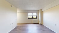 This single-floor unit offers a practical and well-organized layout designed to maximize the use of its available space. The main living area is the centerpiece of the unit, featuring smooth hardwood-style laminate flooring in a warm grey-brown tone that runs throughout. Neutral cream-colored walls create a bright and airy feel, complemented by a textured popcorn ceiling. A large double window fitted with horizontal blinds allows natural light to fill the room while framing a pleasant outdoor view of surrounding greenery. A built-in baseboard heating unit sits beneath the window, ensuring year-round comfort. The open floor plan provides flexible arrangement possibilities for living and dining functions. The dollhouse view reveals a compact but efficient kitchen tucked toward the entry side of the unit, along with a corridor leading to the bathroom. The bathroom features full-height light blue square ceramic wall tiles surrounding a soaking bathtub with a handheld showerhead, a white vanity with a single basin sink, a mirrored medicine cabinet, and classic mosaic tile flooring. The overall ambiance of the unit is clean, functional, and move-in ready, with a timeless design that suits a variety of lifestyles.
