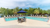 Located at 300 Caughman Farm Ln in Lexington, South Carolina, this two-story community amenity building is designed to offer residents a comfortable and inviting outdoor retreat. The centerpiece of the space is a large, L-shaped outdoor swimming pool with clear blue water, surrounded by an expansive concrete sundeck lined with white lounge chairs — ideal for relaxing in the South Carolina sunshine. A fenced perimeter ensures safety, and lush mature trees and palm trees frame the backdrop, providing a sense of natural privacy and shade. Adjacent to the pool, a covered brick porch with white columns offers a shaded transition area, complete with a package or mailbox station. An outdoor lounge area features comfortable seating with a sofa set, a grill station, and umbrella-shaded dining tables, making it well-suited for casual gatherings. The brick exterior of the amenity building complements the overall traditional architectural style, while the two-story residential buildings visible nearby reflect the broader community setting. This outdoor amenity space combines functional design with a welcoming atmosphere, providing residents with ample space for both relaxation and socializing throughout the year.