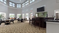 Located at 8600 Cobblefield Drive in Columbia, Maryland, this two-story community clubhouse offers a well-appointed gathering space designed for comfort and functionality. The centerpiece of the main level is a grand lounge area distinguished by dramatic double-height ceilings and a striking clerestory window arrangement that floods the room with natural light. A decorative patterned carpet in earthy tones anchors the space, complementing the neutral gray walls and crisp white trim throughout. The lounge is furnished with clusters of dark-framed seating and round tables, creating an inviting atmosphere for socializing. A built-in bar with dark cabinetry, granite countertops, and bar-height seating serves as both a functional and visual focal point, with a wall-mounted flat-screen television above. Adjacent to the bar, a fully equipped kitchen features stainless steel appliances, granite countertops, dark wood cabinetry, and a glass-canopy range hood. A dedicated fitness center with rubber flooring, exercise equipment, mirrored walls, and vibrant teal and orange accent walls adds an energetic contrast to the more refined lounge aesthetic. Large windows throughout the fitness area frame views of lush surrounding greenery. Connecting corridors with glass-paneled doors and a staircase provide seamless access between levels, completing this versatile and well-designed community facility.