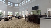 Located at 8600 Cobblefield Drive in Columbia, Maryland, this two-story community clubhouse offers a well-appointed gathering space designed for comfort and functionality. The centerpiece of the main level is a grand lounge area distinguished by dramatic double-height ceilings and a striking clerestory window arrangement that floods the room with natural light. A decorative patterned carpet in earthy tones anchors the space, complementing the neutral gray walls and crisp white trim throughout. The lounge is furnished with clusters of dark-framed seating and round tables, creating an inviting atmosphere for socializing. A built-in bar with dark cabinetry, granite countertops, and bar-height seating serves as both a functional and visual focal point, with a wall-mounted flat-screen television above. Adjacent to the bar, a fully equipped kitchen features stainless steel appliances, granite countertops, dark wood cabinetry, and a glass-canopy range hood. A dedicated fitness center with rubber flooring, exercise equipment, mirrored walls, and vibrant teal and orange accent walls adds an energetic contrast to the more refined lounge aesthetic. Large windows throughout the fitness area frame views of lush surrounding greenery. Connecting corridors with glass-paneled doors and a staircase provide seamless access between levels, completing this versatile and well-designed community facility.
