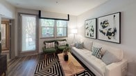 Located in the Natomas Park neighborhood of Sacramento, CA, this single-floor residence offers a thoughtfully designed layout with contemporary finishes throughout. The kitchen is a standout feature, showcasing dark upper cabinetry contrasted by white lower cabinets, stainless steel appliances including a French door refrigerator, a classic white subway tile backsplash, dark countertops, and wood-look flooring — creating a sleek, modern aesthetic. A bar-height dining table with industrial-style stools provides a casual dining option within the kitchen space. The living area is bright and welcoming, with large windows that frame lush outdoor greenery and allow generous natural light to fill the room. It comfortably accommodates a seating arrangement anchored by a bold geometric area rug and is accented by contemporary artwork. The bedroom is a serene retreat, featuring plush carpeting, large windows dressed with elegant patterned curtains, a sage green accent wall, and a walk-in closet with built-in shelving. The bathroom is finished with wood-look flooring, a wide vanity with dark countertops, white shaker-style cabinetry, and a large mirror — combining functionality with clean, modern design.