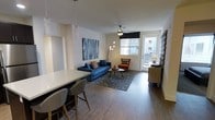This well-designed single-floor space offers a seamless open-concept layout that connects the kitchen and living areas into one cohesive, functional environment. The kitchen is outfitted with dark cabinetry, a stainless steel refrigerator, and a clean white-topped island with bar seating for two, making it ideal for casual dining and entertaining. Pendant lighting with crystal-style fixtures hangs above the island, adding a refined decorative touch.

The living area flows naturally from the kitchen and is anchored by rich wood-look flooring that runs throughout the space. A tufted blue sofa, a patterned area rug, and an accent chair in warm orange tones create a vibrant yet balanced aesthetic. A bold abstract wall canvas serves as a focal point, complementing the modern décor. A ceiling fan with a sleek profile provides both comfort and style overhead.

Natural light filters in through a window fitted with a roman shade, and a glass-paneled exterior door opens to allow additional brightness and ventilation. The overall ambiance is contemporary and inviting, with thoughtful design choices that maximize both form and function throughout the space.