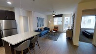 This well-designed single-floor space offers a seamless open-concept layout that connects the kitchen and living areas into one cohesive, functional environment. The kitchen is outfitted with dark cabinetry, a stainless steel refrigerator, and a clean white-topped island with bar seating for two, making it ideal for casual dining and entertaining. Pendant lighting with crystal-style fixtures hangs above the island, adding a refined decorative touch.

The living area flows naturally from the kitchen and is anchored by rich wood-look flooring that runs throughout the space. A tufted blue sofa, a patterned area rug, and an accent chair in warm orange tones create a vibrant yet balanced aesthetic. A bold abstract wall canvas serves as a focal point, complementing the modern décor. A ceiling fan with a sleek profile provides both comfort and style overhead.

Natural light filters in through a window fitted with a roman shade, and a glass-paneled exterior door opens to allow additional brightness and ventilation. The overall ambiance is contemporary and inviting, with thoughtful design choices that maximize both form and function throughout the space.