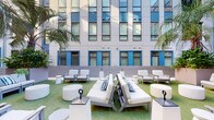 Located on the second floor of a modern building in New Orleans, LA, this open-air garden courtyard offers a beautifully designed outdoor retreat that blends contemporary architecture with lush, tropical landscaping. The courtyard is framed by a multi-story façade featuring a striking combination of neutral panels and bold blue-trimmed windows with horizontal shutters, lending the space a clean, modern aesthetic with a pop of color.

The main gathering area is anchored by artificial turf, which provides a verdant, low-maintenance ground surface across the entire courtyard. Multiple seating arrangements are thoughtfully positioned throughout, featuring light gray upholstered sofas, lounge chairs, and accent tables that invite relaxed conversation and social gatherings. Cylindrical white ottomans and sleek side tables complement the contemporary outdoor furniture, while decorative torch-style lanterns add ambient character.

Tall palm trees and generous planter beds filled with tropical foliage line the perimeter, creating a sense of privacy and a lush, garden-like atmosphere. A wood-paneled raised deck runs along one side of the courtyard, adding warmth and dimension to the space, with additional seating areas featuring teak furniture and festive red chairs. The open-sky setting allows abundant natural light to flood the courtyard, making it an inviting urban oasis in the heart of New Orleans.