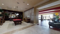This single-floor amenity facility is designed to offer residents a comprehensive recreational and social experience within a refined, elegantly finished environment. The space is anchored by a welcoming lobby featuring large-format travertine tile flooring, crown molding, a wall-mounted flat-screen television, decorative artwork, and glass-paneled entry doors that invite natural light from the outdoors.

Adjacent to the lobby, the game room is a standout social hub appointed with rich dark wood cabinetry, pendant lighting, and a full-size billiards table with a vibrant red felt surface. A velvet settee with decorative pillows adds a comfortable lounge element, while floor-to-ceiling sheer drapery softens the space. Bar-height tables and stools, alongside large framed mirrors, lend a sophisticated, club-like atmosphere.

The fitness center is a well-equipped, generously sized area highlighted by bold red accent walls and durable rubberized flooring. It features a broad selection of strength-training machines, free weights, and cardio equipment including treadmills, ellipticals, and a stair climber. A dedicated stretching and functional training zone houses stability balls, resistance bands, and yoga mats. Large windows throughout the fitness area provide views of the outdoor pool and surrounding greenery, filling the space with natural light.