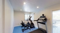 This single-floor fitness space is thoughtfully designed to serve as a dedicated home workout area. The room features a clean, minimalist aesthetic with smooth white walls and a lightly textured ceiling outfitted with recessed lighting, ensuring bright and even illumination throughout. A window fitted with vertical blinds allows natural light to filter in while maintaining privacy, adding a pleasant sense of openness to the space. The workout area is anchored by a protective blue exercise mat that covers the flooring beneath the equipment, providing both safety and a defined workout zone. The room is equipped with a treadmill, an elliptical trainer, and a stationary bike, offering a well-rounded selection of cardiovascular equipment suited to a variety of fitness routines. The layout is efficient and uncluttered, allowing for comfortable movement between machines. The overall ambiance is functional and focused, making it an inviting environment for daily exercise. Whether used for high-intensity cardio sessions or low-impact recovery workouts, this fitness room delivers a practical and purpose-built space for maintaining an active lifestyle.