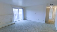 This single-floor space features a bright, open living area finished with wall-to-wall carpeting in a neutral tone, contributing to a clean and understated aesthetic. The room is defined by its simplicity, with smooth white walls and a plain ceiling that together create a bright, airy atmosphere. A sliding glass door serves as a central architectural feature, filling the space with natural light and providing direct access to an outdoor balcony enclosed by a white railing. The view through the glass door reveals surrounding trees and a neighboring structure, offering a sense of connectivity to the outdoors. Electrical outlets are evenly distributed along the walls, reflecting practical functionality throughout. The overall ambiance of the space is calm and neutral, providing a versatile blank canvas well-suited for a variety of layout and décor configurations.
