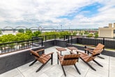 This expansive rooftop terrace is a thoughtfully designed outdoor destination offering a variety of distinct zones for relaxation, dining, and socializing. The entire space is finished with large-format light concrete tiles, providing a clean, modern foundation that ties the various areas together cohesively.

Multiple pergola structures with warm wood slat ceilings and bold dark steel frames define key gathering spaces throughout the terrace. String lights beneath the pergolas add an inviting ambiance, while an outdoor mounted television creates a comfortable media lounge area furnished with plush gray sectional sofas and lounge chairs.

Elevated bar-height tables and chairs are arranged beneath a pergola for open-air dining, perfectly positioned to take advantage of the sweeping views. A separate lounge corner features Adirondack-style wooden chairs paired with small bistro tables, ideal for casual conversation. Additional seating groupings with modern upholstered outdoor furniture are distributed across the terrace to accommodate a variety of uses.

The terrace is enclosed by dark metal railing, framing dramatic panoramic views of a wide river, a signature arched bridge, a Ferris wheel, and a prominent downtown skyline with a distinctive historic tower. This rooftop space excels as a venue for both everyday use and special gatherings, combining modern industrial design with an unmatched outdoor urban setting.