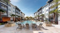This expansive outdoor amenity space is designed to evoke a resort-like atmosphere, centered around a generously sized swimming pool that stretches the length of the courtyard. The pool deck is finished in light-toned travertine-style pavers and lined with contemporary chaise lounges and accent tables, providing ample space for sunbathing and relaxation. Flanking the pool on both sides are multi-story residential buildings with modern architectural detailing, featuring a mix of gray and white facades, balconies, and large windows that overlook the amenity area.

Lush artificial turf lawns border the pool deck, offering a soft, green counterpoint to the hard-surface paving. These lawn areas serve as informal recreation zones, complete with cornhole boards and sculptural seating elements. Mature palm trees are planted throughout the space, reinforcing the tropical ambiance and providing natural shade.

A warm-toned wood pergola structure anchors one end of the courtyard, fitted with string lights overhead and furnished with bistro-style tables and teal chairs. Cedar privacy screening adds definition to this intimate seating zone. The entire space benefits from abundant natural light and open sky views, creating a vibrant, social outdoor environment suited for both leisure and community gatherings throughout the day.