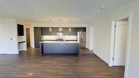 This single-floor space showcases a contemporary open-concept design that seamlessly blends style and functionality. The kitchen serves as the heart of the space, featuring dark, flat-panel cabinetry that extends to the ceiling for generous storage capacity. A large central island with a crisp white countertop provides ample workspace and doubles as a casual dining or gathering spot. Three elegant pendant lights hang above the island, casting a warm, inviting glow over the area. The kitchen is equipped with stainless steel appliances, including a full-size refrigerator, and is finished with a tasteful tile backsplash that adds texture and visual interest. Recessed lighting throughout the ceiling ensures the space is well-lit and bright. Rich, dark hardwood flooring flows continuously across the open layout, grounding the space with warmth and depth. The clean white walls and crisp trim provide a neutral backdrop that highlights the contrast between the dark cabinetry and light countertops. The open floor plan allows the kitchen to flow naturally into the adjoining living areas, creating a spacious and cohesive environment that is both modern and welcoming.