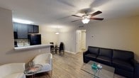 This single-floor unit showcases an open-concept layout that seamlessly connects the living, dining, and kitchen areas into one cohesive space. The living area is anchored by a ceiling fan with integrated lighting, which adds both functionality and a focal point overhead. A textured ceiling runs throughout, complementing the neutral, light-toned walls that create a clean and welcoming atmosphere.

The flooring is finished in wood-style planks, lending warmth and continuity across the shared living spaces. The living area comfortably accommodates a sofa and accent chair arrangement centered around a glass coffee table, while the dining area features a compact table and chair set illuminated by a pendant light fixture.

The kitchen is partially visible beyond a raised breakfast bar counter, which provides a natural dividing element while maintaining the open feel. Dark cabinetry and a black refrigerator offer a contrasting design detail. A hallway visible toward the rear of the space leads to additional rooms, suggesting a well-organized and functional floor plan. A programmable thermostat is mounted on the wall, indicating modern climate control features throughout.