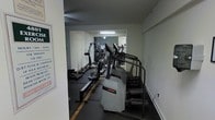 This single-floor fitness room offers a focused workout environment outfitted with a selection of cardio equipment to support a range of exercise routines. The room features dark rubber flooring, which provides both durability and a non-slip surface suited for high-traffic use. A large mirror spans the rear wall, visually expanding the space and allowing users to monitor their form during workouts. The equipment lineup includes a Life Fitness stair climber, an elliptical trainer, a rowing machine, and additional cardio machines positioned along the length of the room. Overhead fluorescent lighting provides functional illumination throughout. The white walls and clean layout contribute to a straightforward, no-frills atmosphere designed with utility in mind. The arrangement of machines along the perimeter maximizes usable floor space, making the room efficient despite its modest footprint. Overall, this fitness room serves as a practical and well-equipped space for cardiovascular training.