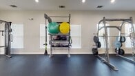 Located at Integra Springs in Kannapolis, North Carolina, this well-appointed fitness center offers a comprehensive workout environment designed to support a wide range of exercise routines. The single-floor facility features durable dark rubber flooring throughout, providing a clean and functional surface suited for both cardio and strength training activities.

The cardio zone is equipped with multiple treadmills, elliptical machines, and stationary bikes, positioned along windowed walls fitted with slatted blinds that allow natural light to filter into the space. Ceiling fans installed throughout the room enhance air circulation and comfort during workouts.

The strength training area is anchored by a full rack of dumbbells spanning a wide range of weights, accompanied by a flat bench for versatile exercise options. A dedicated functional training station holds stability balls, medicine balls, kettlebells, resistance bands, and TRX suspension trainers, while an adjacent weight plate rack supports barbell training needs.

Large wall-mounted mirrors along one side of the room visually expand the space and allow users to monitor their form during exercise. A wall-mounted flat-screen television adds an entertainment option for cardio sessions. The neutral white walls and recessed lighting create a bright, clean atmosphere, while a glass-paned entry door adds an architectural touch that ties the space together.