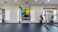 Located at Integra Springs in Kannapolis, North Carolina, this well-appointed fitness center offers a comprehensive workout environment designed to support a wide range of exercise routines. The single-floor facility features durable dark rubber flooring throughout, providing a clean and functional surface suited for both cardio and strength training activities.

The cardio zone is equipped with multiple treadmills, elliptical machines, and stationary bikes, positioned along windowed walls fitted with slatted blinds that allow natural light to filter into the space. Ceiling fans installed throughout the room enhance air circulation and comfort during workouts.

The strength training area is anchored by a full rack of dumbbells spanning a wide range of weights, accompanied by a flat bench for versatile exercise options. A dedicated functional training station holds stability balls, medicine balls, kettlebells, resistance bands, and TRX suspension trainers, while an adjacent weight plate rack supports barbell training needs.

Large wall-mounted mirrors along one side of the room visually expand the space and allow users to monitor their form during exercise. A wall-mounted flat-screen television adds an entertainment option for cardio sessions. The neutral white walls and recessed lighting create a bright, clean atmosphere, while a glass-paned entry door adds an architectural touch that ties the space together.