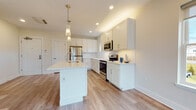 This single-floor space presents a clean, contemporary design highlighted by light hardwood-style flooring and neutral warm-toned walls that carry consistently throughout. The open-concept kitchen serves as a central feature, equipped with white shaker-style cabinetry, light quartz countertops, a subway tile backsplash, and a full suite of stainless steel appliances including a refrigerator, range, and microwave. A centrally positioned kitchen island with an integrated sink provides additional workspace and storage, while elegant glass pendant lights overhead add a refined touch. Recessed lighting illuminates the kitchen and adjoining living area, which benefits from multiple large windows that invite generous natural light and outdoor views.

The living space is notably open and versatile, with room for various furniture configurations. A wall-mounted mini-split unit provides climate control. Two bathrooms serve the unit: a full bathroom featuring a white shaker vanity, LED-backlit mirror, and a tub with marble-style surround; and a primary bathroom with a double-sink vanity, dual framed mirrors, and a glass-enclosed shower with marble-style tile. Additional bedrooms offer clean, well-proportioned spaces with the same consistent flooring and neutral palette found throughout.