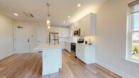 This single-floor space presents a clean, contemporary design highlighted by light hardwood-style flooring and neutral warm-toned walls that carry consistently throughout. The open-concept kitchen serves as a central feature, equipped with white shaker-style cabinetry, light quartz countertops, a subway tile backsplash, and a full suite of stainless steel appliances including a refrigerator, range, and microwave. A centrally positioned kitchen island with an integrated sink provides additional workspace and storage, while elegant glass pendant lights overhead add a refined touch. Recessed lighting illuminates the kitchen and adjoining living area, which benefits from multiple large windows that invite generous natural light and outdoor views.

The living space is notably open and versatile, with room for various furniture configurations. A wall-mounted mini-split unit provides climate control. Two bathrooms serve the unit: a full bathroom featuring a white shaker vanity, LED-backlit mirror, and a tub with marble-style surround; and a primary bathroom with a double-sink vanity, dual framed mirrors, and a glass-enclosed shower with marble-style tile. Additional bedrooms offer clean, well-proportioned spaces with the same consistent flooring and neutral palette found throughout.